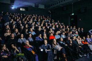 Pełna sala kinowa – dziesiątki osób siedzących w rzędach ciemnej sali, patrzących w stronę ekranu. Widać światło projektora z tyłu, różne reakcje na twarzach widzów, a całość jest oświetlona przytłumionym, niebieskawym światłem.